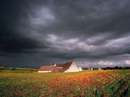 A farmhouse under a dramatic, cloudy sky sits in a colorful field, with storm clouds looming above, creating a striking contrast.