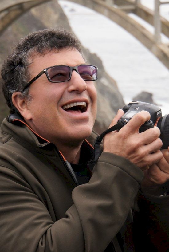 A person is joyfully holding a camera near a scenic bridge and natural landscape.