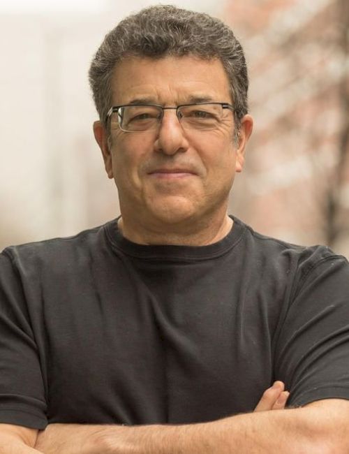 A person wearing a black shirt stands outside with arms crossed, smiling slightly, and buildings blurred in the background.