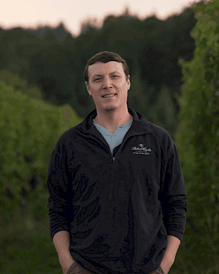 A person stands outdoors in a vineyard setting, wearing casual clothing, with trees in the background.