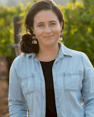 A person in a denim shirt stands in front of greenery, possibly a vineyard, with a warm background light ending the sentence.