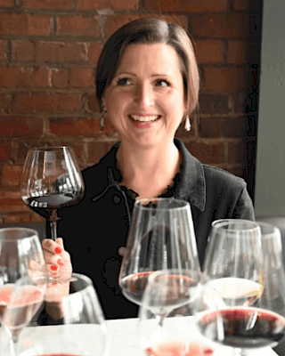 A person is surrounded by various wine glasses, smiling and holding one glass up.