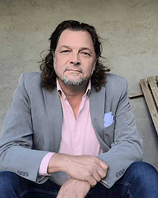 A man with long hair, wearing a gray blazer and pink shirt, sits with his hands clasped in front of a neutral background.