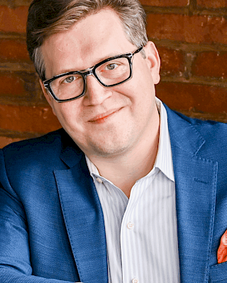 A person wearing glasses and a blue suit jacket is sitting in front of a brick wall, smiling at the camera.