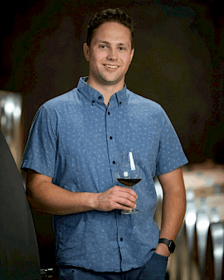 A person in a blue shirt holds a wine glass, standing indoors with a blurred background.