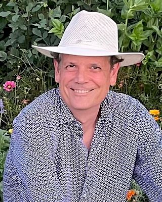 A person wearing a hat and patterned shirt smiles while sitting in a garden with green plants and colorful flowers.