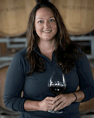 A person is holding a glass of red wine in front of wine barrels.