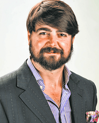 A person with a beard and styled hair, wearing a dark blazer, striped shirt, and a pocket square, smiling against a light background.