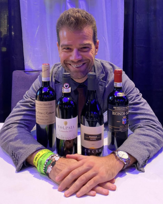 A man is sitting at a table with four bottles of wine in front of him, wearing a suit jacket and colorful wristbands.