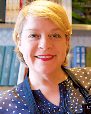 A person smiling in front of bookshelves, wearing a polka-dot blouse and an apron, with short blonde hair.