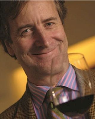 A person smiling while holding a glass of red wine. They are wearing a striped shirt and jacket, with a soft background glow.