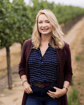 A person standing in a vineyard, smiling, wearing a dark cardigan and blue patterned shirt. The background features rows of grapevines.