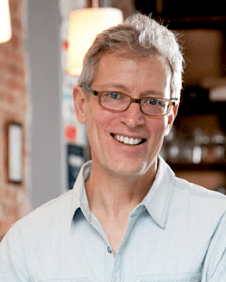 A smiling person with glasses wearing a light shirt stands indoors, with blurred background elements and warm lighting in the scene.