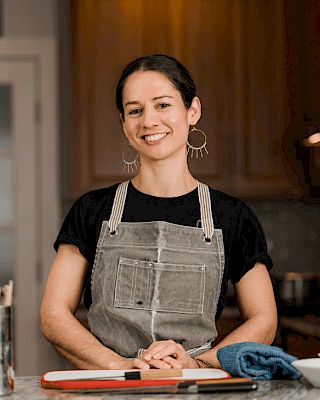 A person in a gray apron smiles in a kitchen setting, standing behind a cutting board with a towel beside it.