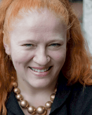 The image shows a person with red hair, smiling at the camera, wearing a necklace with large beads and a dark top, with trees in the background.