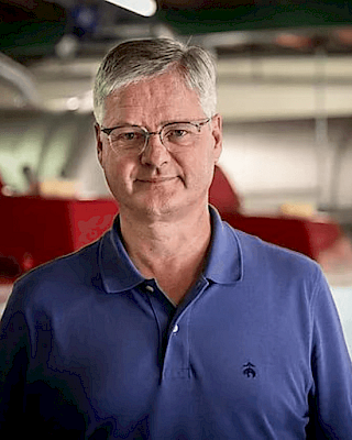 A person wearing glasses and a blue polo shirt stands indoors with industrial equipment blurred in the background.