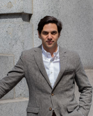 A person in a gray blazer and white shirt stands against a stone wall, hands in pockets, outdoors.