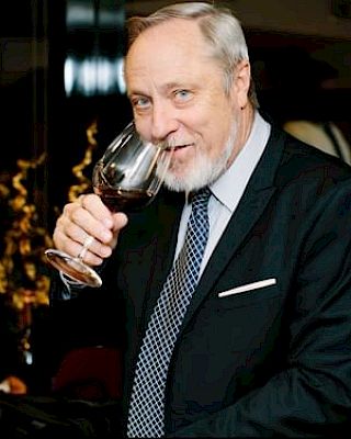 A man in a suit sips wine from a glass, surrounded by large wine bottles and elegant decor in the background.