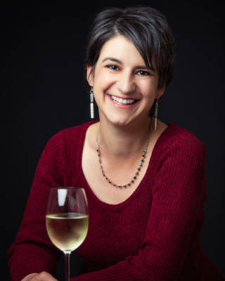 A person with short hair, wearing a red sweater and earrings, smiles while holding a glass of white wine in front of a dark background.