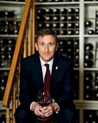 A person in a suit holding a wine glass is sitting on stairs, surrounded by shelves filled with wine bottles in the background.