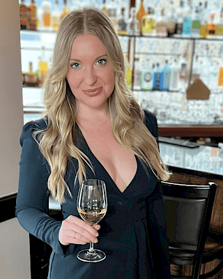 A person with long hair is holding a wine glass, standing in front of a bar with various bottles visible in the background.