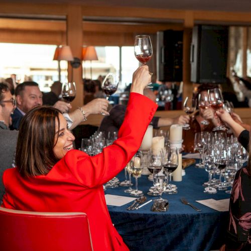 People raising glasses in a toast at a restaurant table, filled with wine glasses and plates. Everyone appears to be in a celebratory mood.