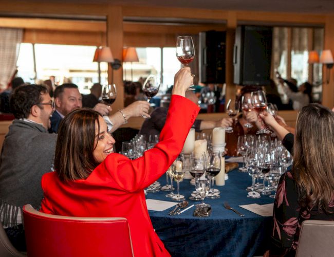 People raising glasses in a toast at a restaurant table, filled with wine glasses and plates. Everyone appears to be in a celebratory mood.