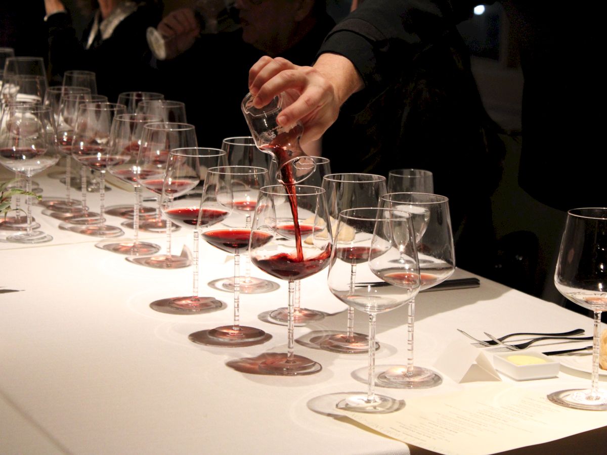 A person is pouring red wine into a row of wine glasses arranged on a table, with several participants possibly involved in a tasting event.