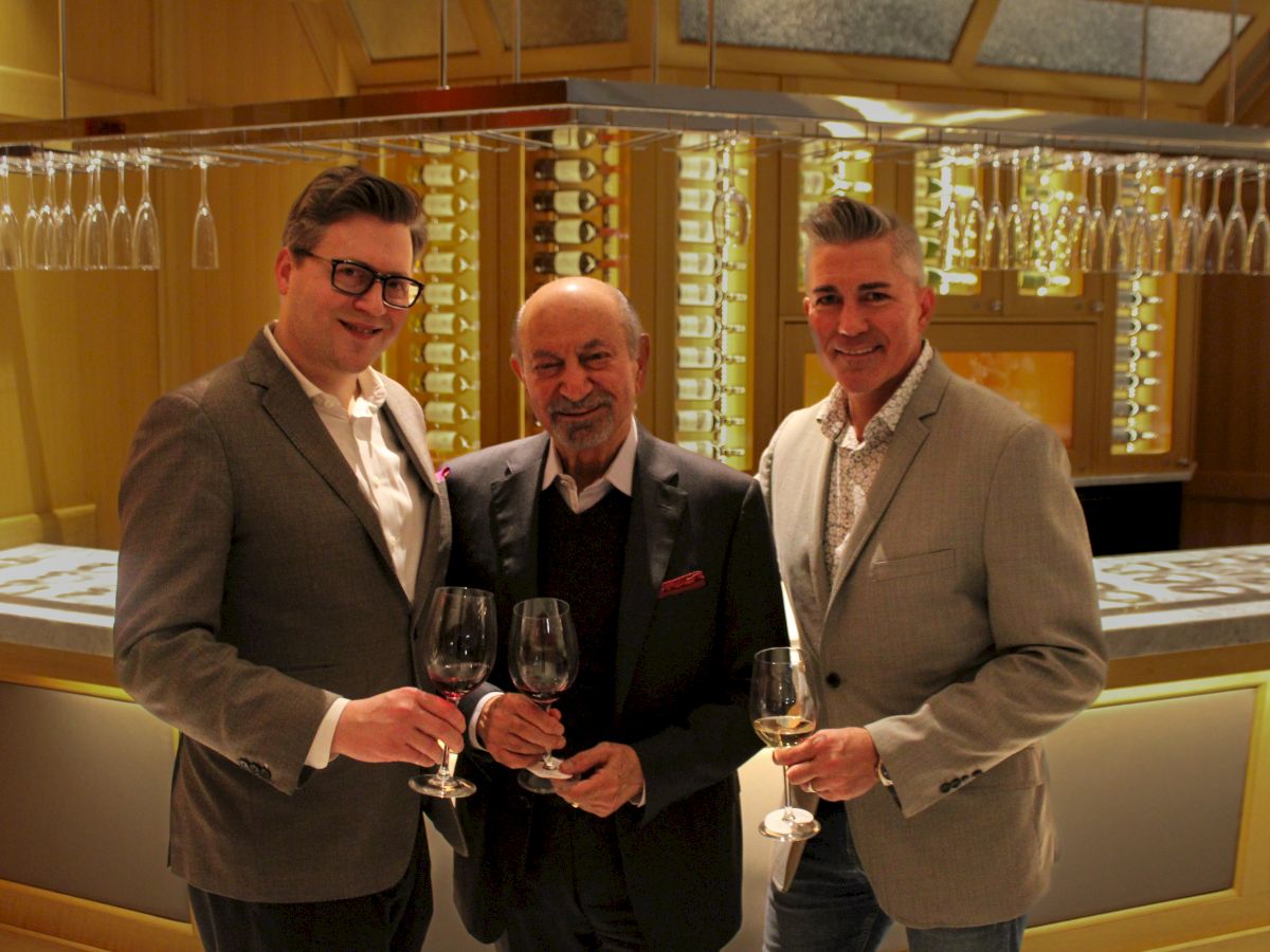 Three men in suits are holding wine glasses, standing in a stylish bar setting with glasses hanging above.