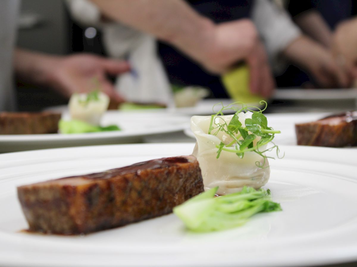 A plated dish featuring a seared meat portion, greens, and a garnish, with chefs preparing food in the background.