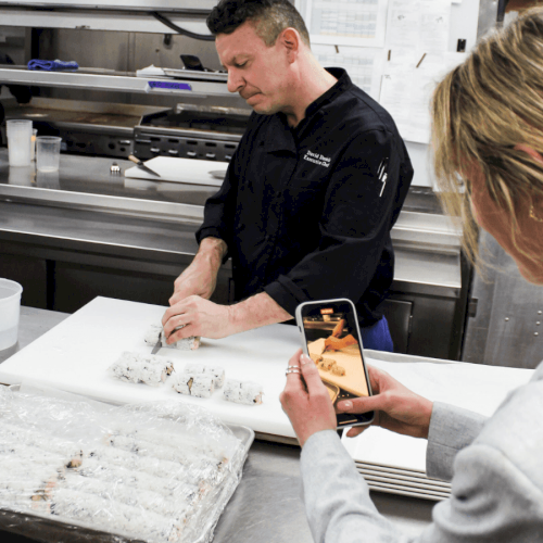 A chef is slicing sushi rolls on a cutting board while someone takes a photo with a smartphone in a kitchen setting.