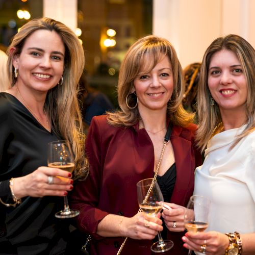 Three women are smiling and holding wine glasses at a social event indoors.