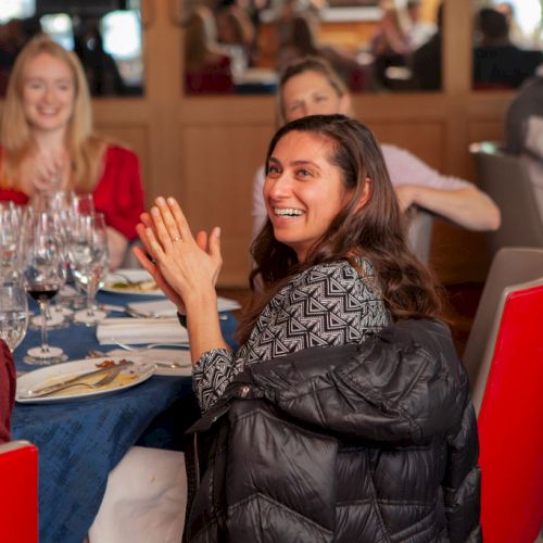 A group of people seated at a restaurant table, applauding and smiling, with wine glasses and plates in front of them.