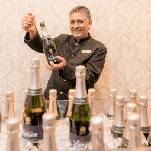 A person in a uniform holds a champagne bottle, standing behind a table with several more bottles on ice and a patterned wallpaper background.