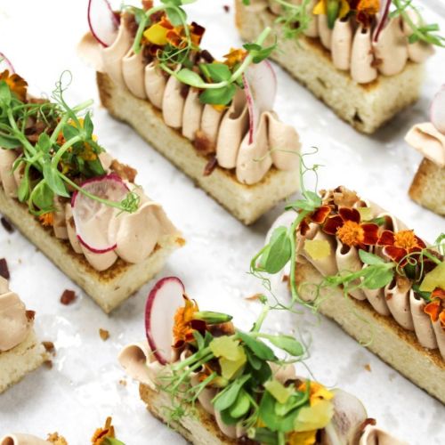 Gourmet appetizers with creamy toppings, edible flowers, and microgreens on bread slices, artistically arranged on a white surface.