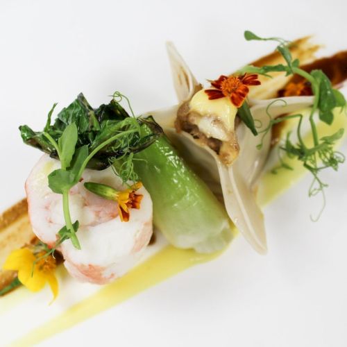 A gourmet dish featuring elegantly presented seafood, vegetables, and garnished with fresh herbs and edible flowers on a white background.