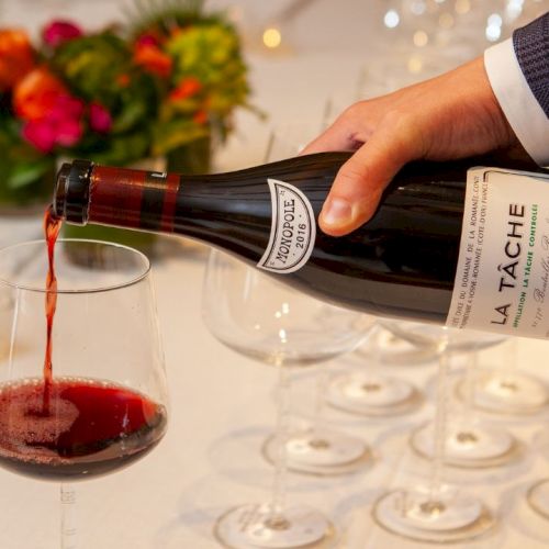 A person pours red wine from a labeled bottle into a glass on a table set with glasses and a floral centerpiece.