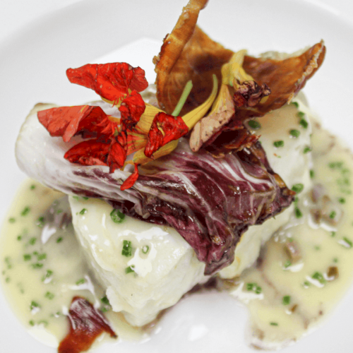 A gourmet dish featuring fish topped with radicchio, mushrooms, herbs, and a creamy sauce, artistically plated.