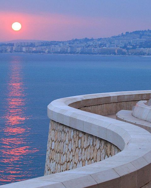 A serene coastal scene at sunset, featuring a curved walkway by the sea and a distant cityscape with a pinkish sky.