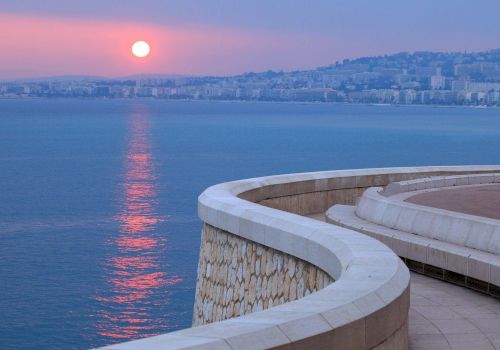 A serene coastal scene at sunset, featuring a curved walkway by the sea and a distant cityscape with a pinkish sky.