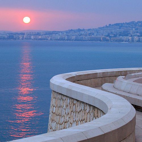 A serene coastal scene at sunset, featuring a curved walkway by the sea and a distant cityscape with a pinkish sky.