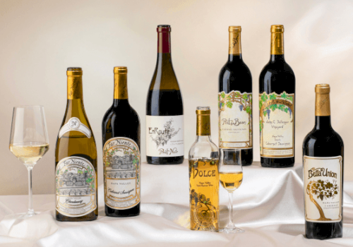 A selection of wine bottles and glasses on a draped surface, showcasing a variety of labels and wine types.