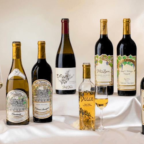 A selection of wine bottles and glasses on a draped surface, showcasing a variety of labels and wine types.