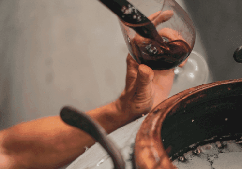 A person is holding a wine glass near a wooden barrel, possibly during a wine tasting or production process.