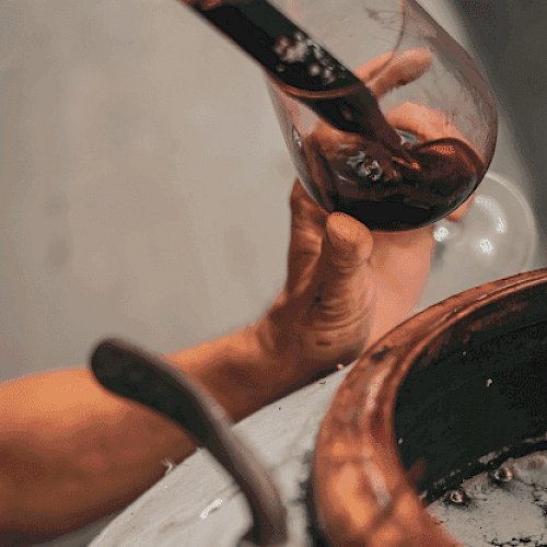 A person is holding a wine glass near a wooden barrel, possibly during a wine tasting or production process.