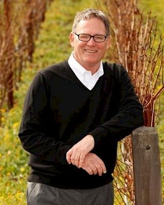 A man in glasses and a black sweater is standing in a vineyard, leaning on a post, with rows of grapevines in the background.