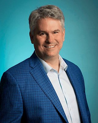 The image shows a man with gray hair wearing a blue checkered suit and white shirt against a teal background, smiling at the camera.