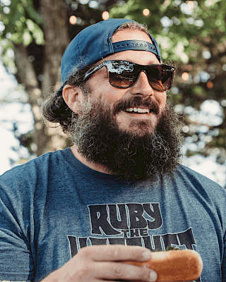 A man with sunglasses and a beard is smiling while holding a hotdog. He's wearing a cap and a T-shirt outdoors.