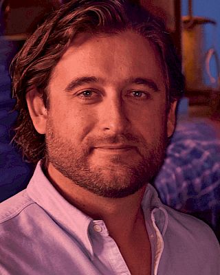 The image shows a person with a beard and wavy hair, wearing a light-colored shirt, standing in an indoor setting.