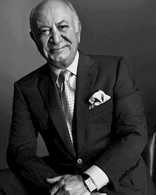 The image shows a man in a suit, sitting and smiling. He has a pocket square, tie, and watch, posing against a plain background.